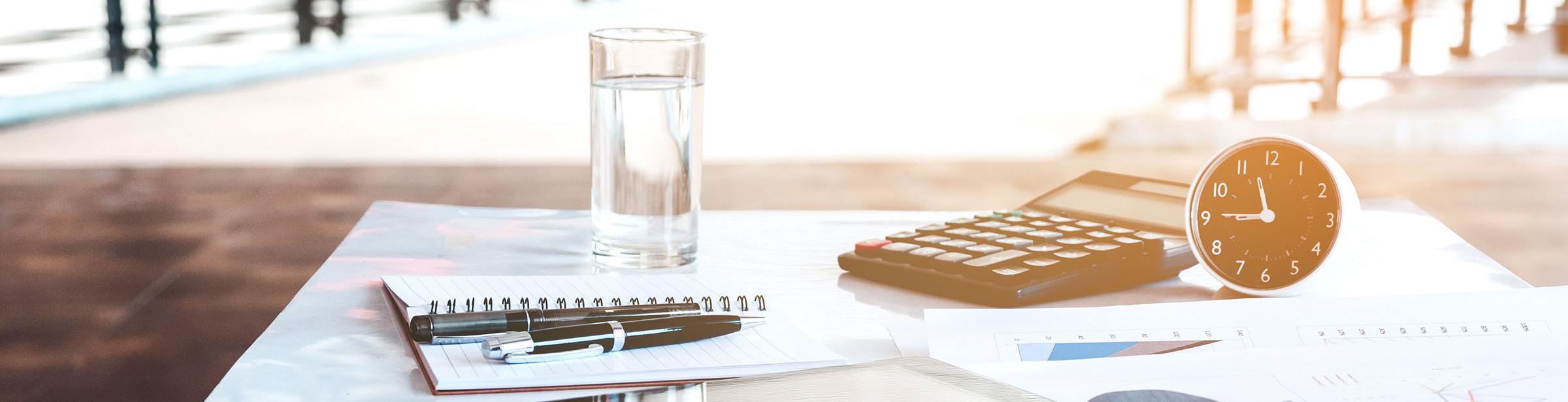glass of water, clock, calculator, day planner and writing pen on a desk